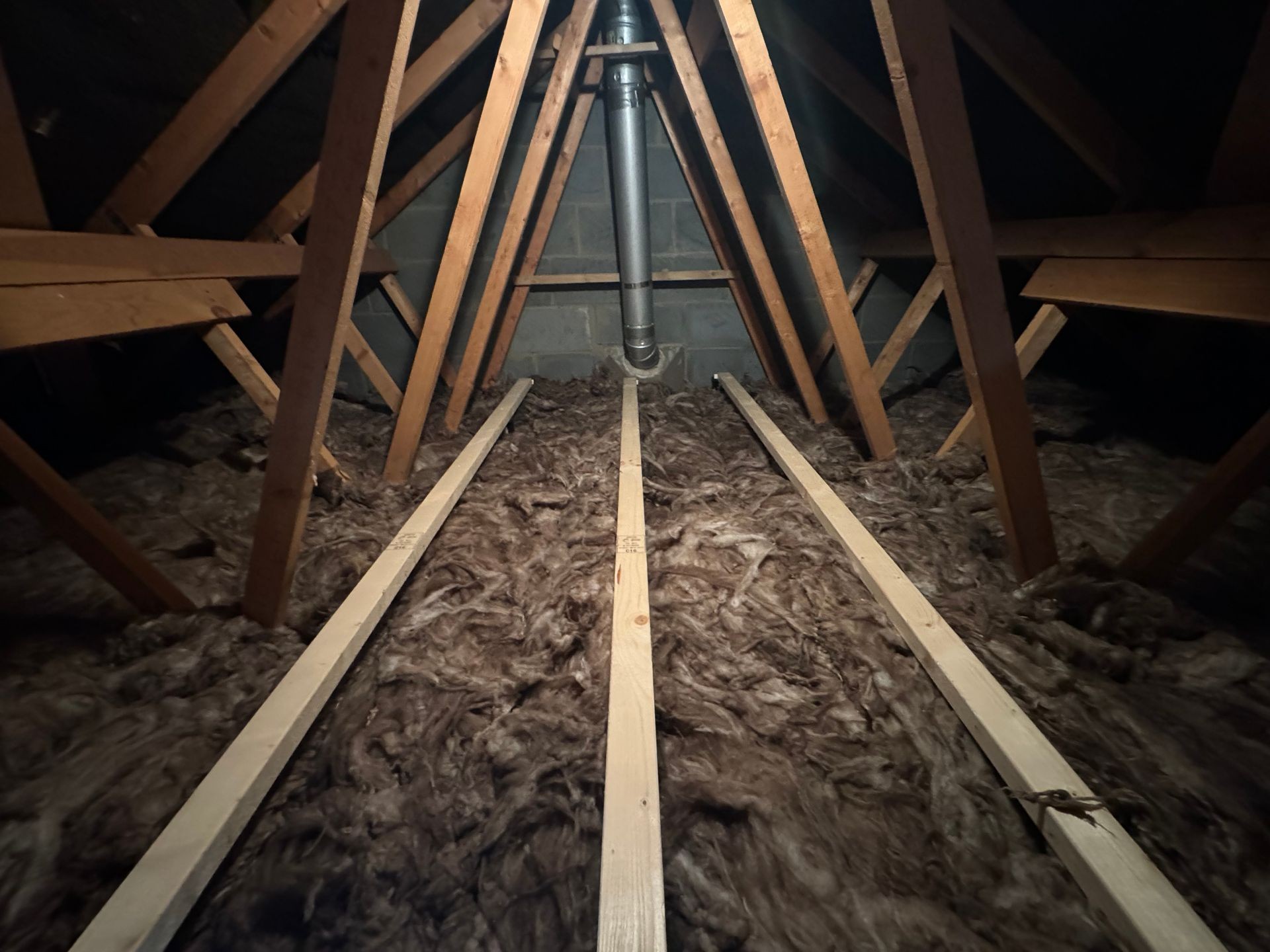 Attic space with wooden beams, insulation on the floor, and a metal pipe running vertically.