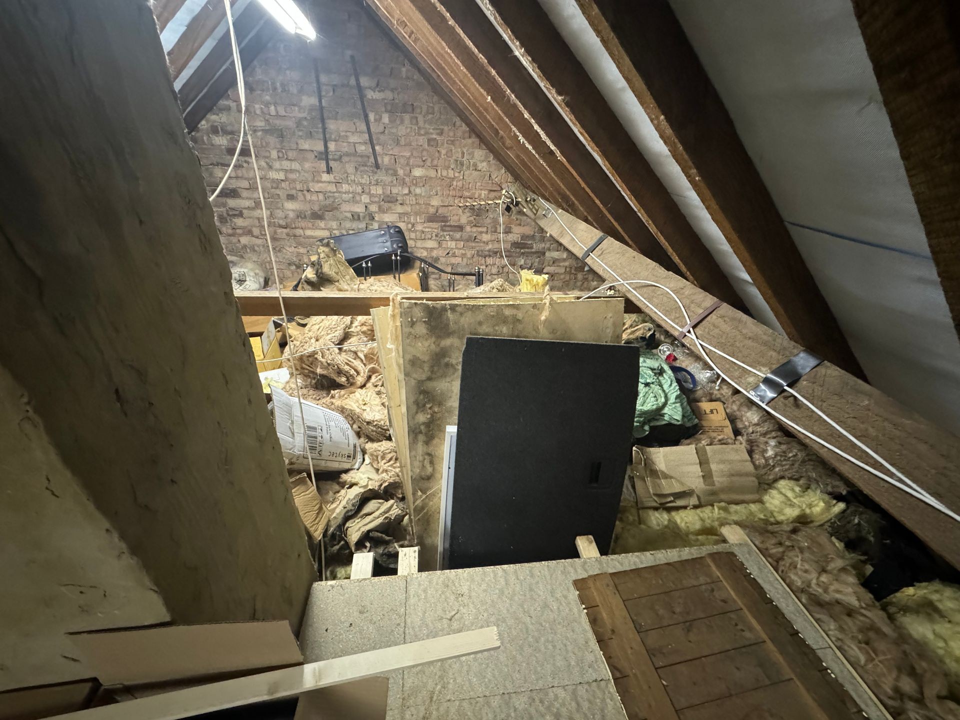 Cluttered attic with insulation, wooden beams, and various stored items.