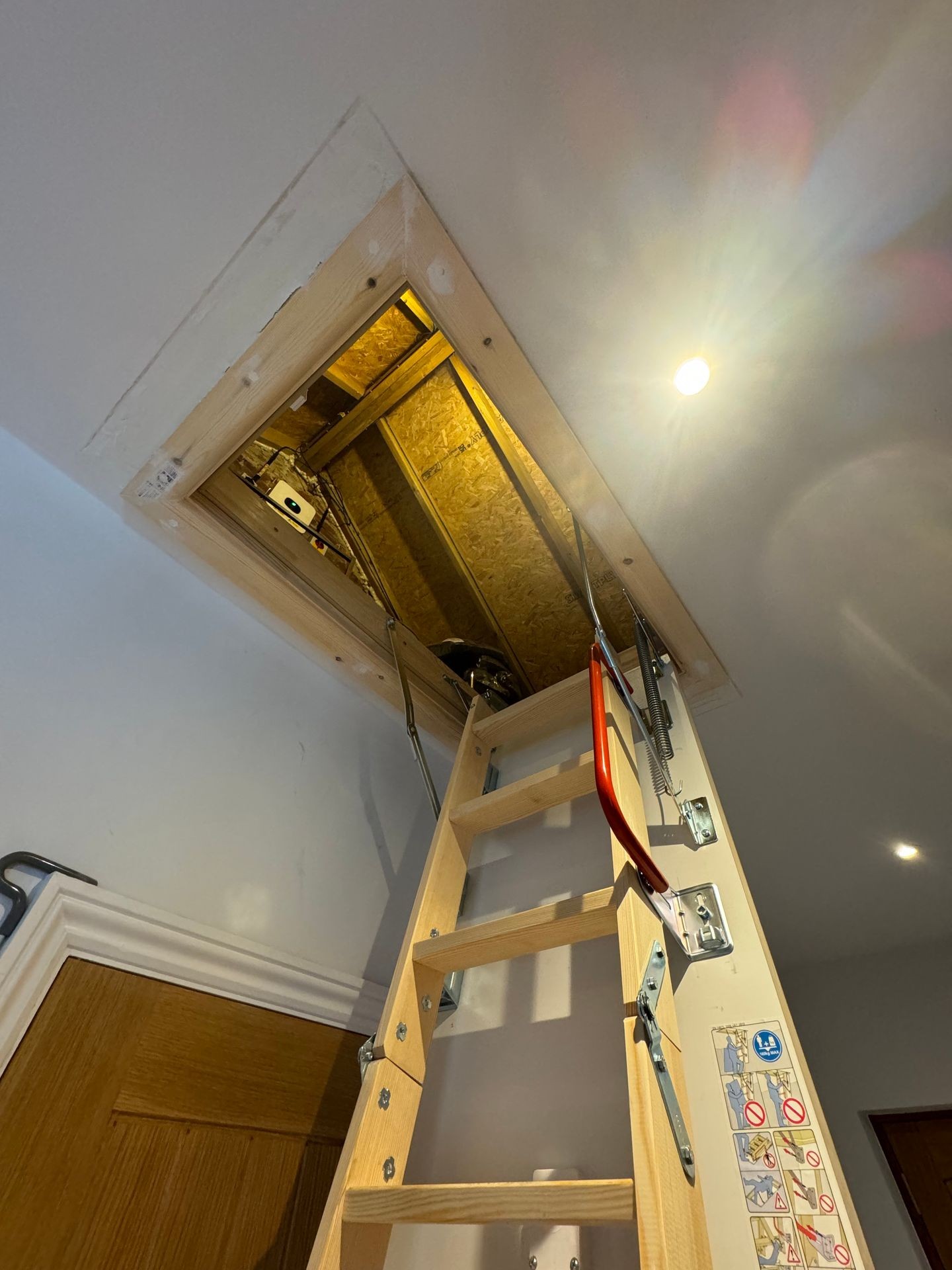 Wooden attic ladder extended from ceiling with visible safety instructions and glowing light bulb nearby.